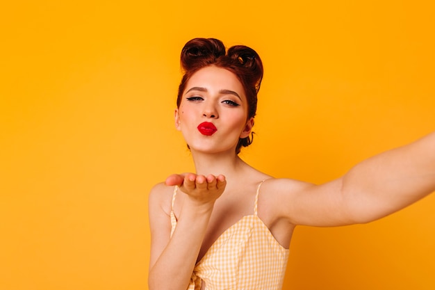 Sinnliche hübsche Frau, die Luftkuss sendet. Schönes Pinup-Mädchen, das Selfie auf gelbem Raum nimmt.