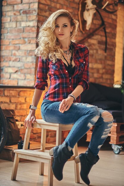 Sinnliches blondes Hipster-Mädchen mit langen lockigen Haaren in Fleece-Shirt und Jeans hält eine Tasse Morgenkaffee auf einem Holzhocker in einem Studio mit Loft-Interieur und blickt in eine Kamera.