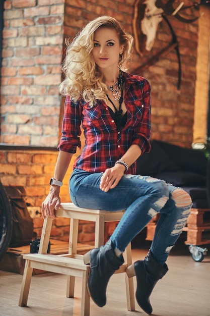 Sinnliches blondes Hipster-Mädchen mit langen lockigen Haaren in Fleece-Shirt und Jeans hält eine Tasse Morgenkaffee auf einem Holzhocker in einem Studio mit Loft-Interieur und blickt in eine Kamera.