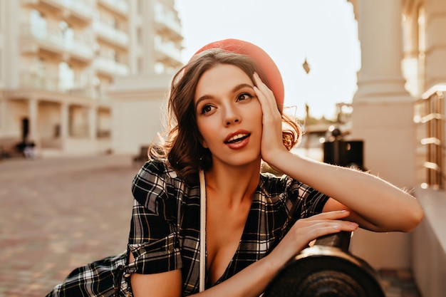Sinnliches französisches Mädchen mit eleganter Frisur, die auf Bank sitzt. Außenporträt der schönen europäischen Frau in der roten Baskenmütze, die auf Stadt aufwirft.
