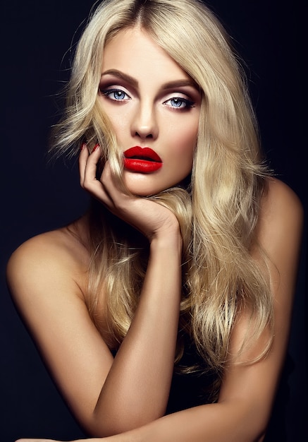 sinnliches Glamourporträt der schönen blonden Frauenmodelldame mit hellem Make-up und roten Lippen, mit gesundem lockigem Haar auf schwarzem Hintergrund