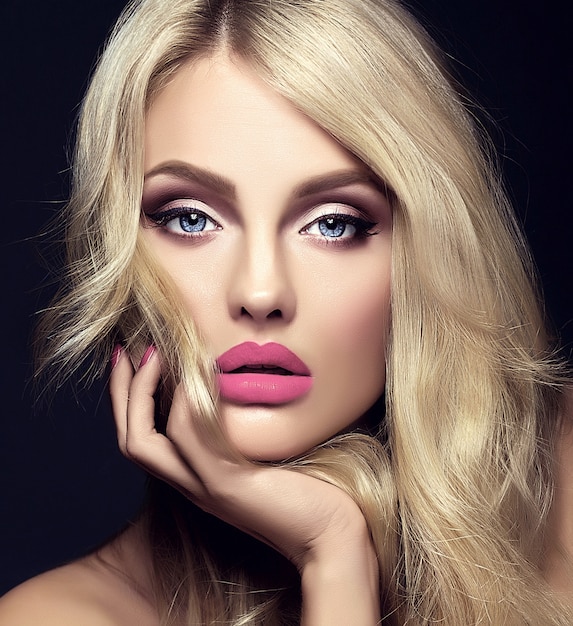 sinnliches Glamourporträt der schönen blonden Modellfrau mit hellem Make-up und roten Lippen, die ihr Gesicht berühren, mit gesundem lockigem Haar auf schwarzem Hintergrund