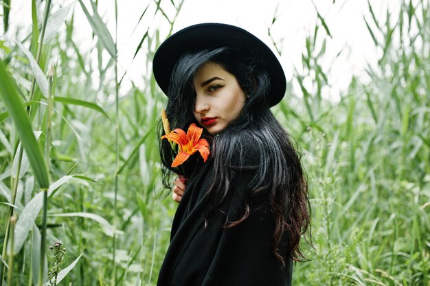 Sinnliches Mädchen ganz in schwarz-roten Lippen und Hut Goth-dramatische Frau hält orangefarbene Lilienblume auf gewöhnlichem Schilf