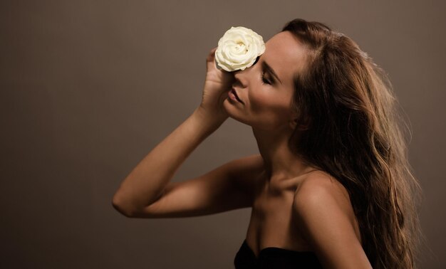 Sinnliches Model, das weiße Rose mit geschlossenen Augen hält Dame mit langen braunen Haaren posiert für Fotografen im Studio
