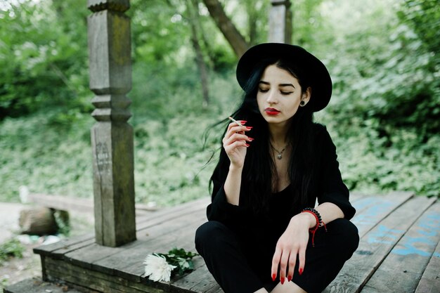 Sinnliches Rauchermädchen ganz in schwarzroten Lippen und Hut Dramatische Goth-Frau, die eine dünne Zigarette raucht