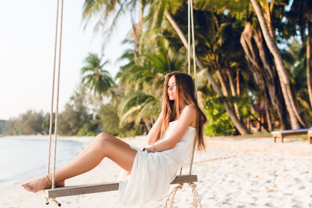 Sinnliches zartes Mädchen, das auf einer Schaukel sitzt, die weißes Kleid trägt. Mädchen hat die Augen geschlossen. Sie hat lange dunkle Haare. Sie hat Armbänder an Arm und Bein. Die Schaukel ist am Strand mit grünen Palmen