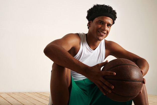 sitzender lächelnder freundlicher afroamerikanischer Basketballspieler mit einem Afro, der weiße und grüne Uniform trägt, die einen braunen Lederball hält