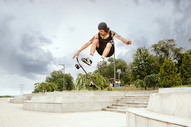 Skateboarder machen an bewölkten Tagen einen Trick auf der Straße der Stadt. Junger Mann in Turnschuhen und Mütze reiten und Longboarding auf dem Asphalt. Konzept von Freizeitbeschäftigung, Sport, Extrem, Hobby und Bewegung.