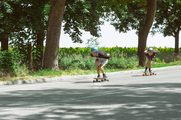 Skateboarder machen an sonnigen Tagen einen Trick auf der Straße der Stadt. Junge Männer in Ausrüstung reiten und Longboarding in der Nähe von Wiese in Aktion. Konzept von Freizeitbeschäftigung, Sport, Extrem, Hobby und Bewegung.