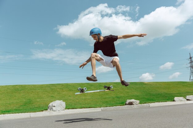 Skateboarder macht an einem sonnigen Tag einen Trick auf der Straße der Stadt. Junger Mann im Ausrüstungsreiten und Longboarden nahe Wiese in Aktion. Konzept von Freizeitbeschäftigung, Sport, Extrem, Hobby und Bewegung.