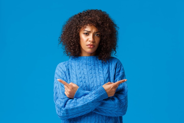 Skeptisch und unsicher, unentschlossen ernsthaft aussehende afroamerikanische Frau mit Afro-Haarschnitt, die die Stirn runzelt, verurteilend und unsicher, mit den Fingern seitwärts, links und rechts auf blauem Hintergrund zeigend.