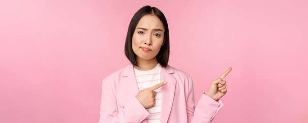 Skeptische asiatische Geschäftsfrau, Verkäuferin, die schmollend und missbilligend aussieht, während sie mit dem Finger nach rechts zeigt und schlechte Informationen zeigt, die beunruhigende Nachrichten über rosa Hintergrund stehen