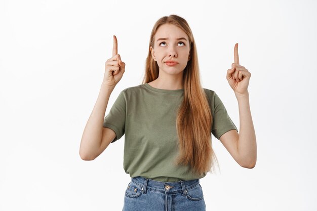Skeptisches blondes Mädchen grinst, zeigt mit den Fingern nach oben und schaut auf das Produktbanner mit verurteilendem unzufriedenem Gesicht, mag den Artikel oben nicht und steht auf weißem Hintergrund