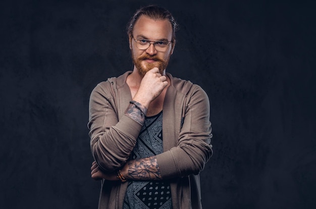 Smarter, nachdenklicher Rotkopf-Hipster mit Vollbart und Brille in Freizeitkleidung, posiert mit der Hand am Kinn in einem Studio. Getrennt auf dem dunklen Hintergrund.