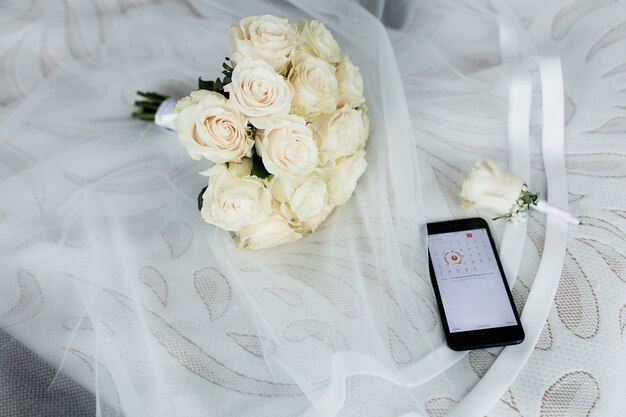 Smartphone mit geöffnetem Kalender, Hochzeitsknopfloch und Hochzeitsstrauß aus weißen Rosen auf dem Schleier