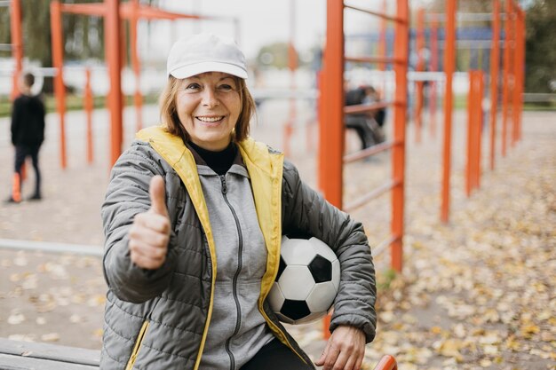 Smiley ältere Frau, die Fußball hält und Daumen aufgibt, während sie trainiert