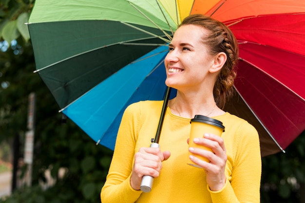 Smiley-Frau, die eine Tasse Kaffee unter einem bunten Regenschirm hält