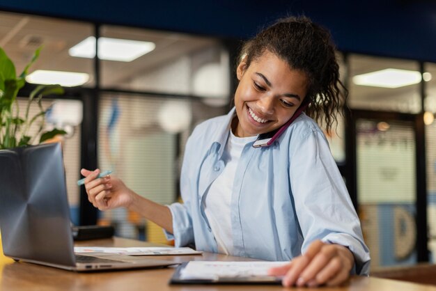 Smiley-Frau im Büro, das Laptop benutzt und auf Smartphone spricht