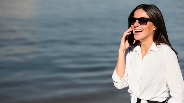 Smiley-Frau mit Sonnenbrille, die am Strand am Telefon spricht