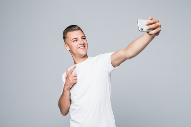 Smiley junger Mann trägt ein weißes T-Shirt und macht ein Selfie mit einem silbernen Smartphone.