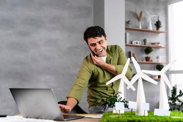 Smiley-Mann, der an einem umweltfreundlichen Windkraftprojekt arbeitet, während er am Telefon spricht und Laptop verwendet