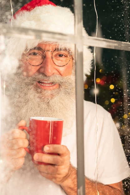 Kostenloses Foto smiley-mann im weihnachtsmannhut, der becher hält und durch fenster schaut