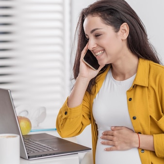 Smiley schwangere frau, die am telefon mit laptop spricht
