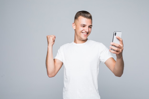 Smiley starker junger Mann trägt ein weißes T-Shirt und macht ein Selfie mit einem silbernen Smartphone.