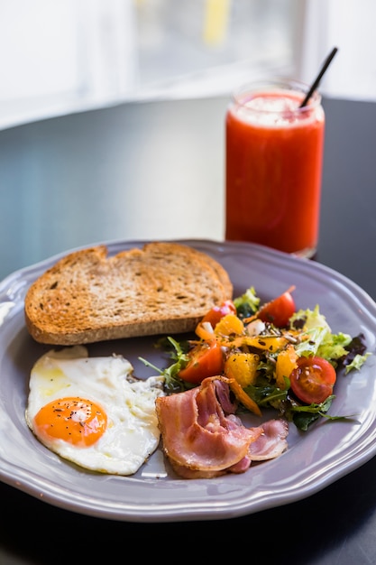 Smoothie im Glas; Toast; Speck; Spiegelei; Salat auf grauer Platte über der schwarzen Tabelle