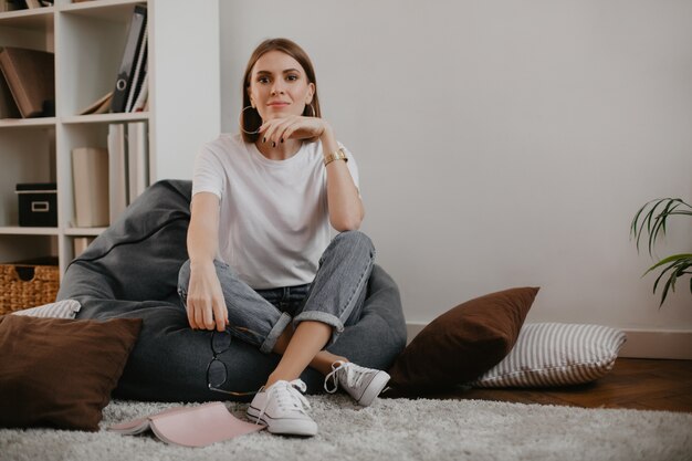 Snapportrait der ruhigen jungen Frau im stilvollen Outfit, das im grauen Taschenstuhl im weißen Raum sitzt.