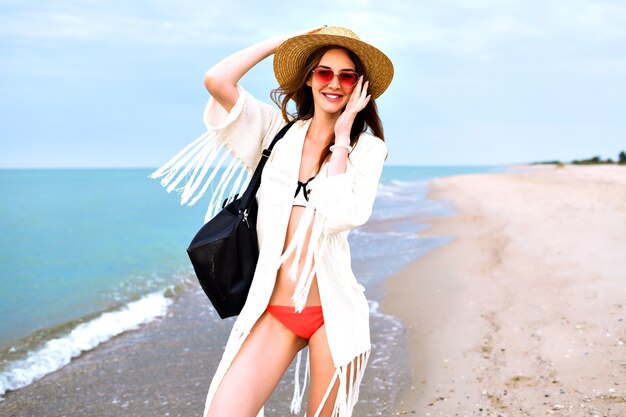 Sommer-Außenporträt der hübschen blonden Frau, die Bikini, Boho-Art-Jacke und Sonnenbrille trägt und nahe Ozean, glückliche Reiseurlaubsstimmung aufwirft.