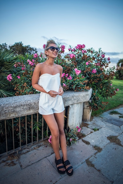 Sommer sonnigen Lebensstil Mode Porträt der jungen stilvollen Hipster Frau sitzt auf Beton, trägt süße trendy weiße Outfit. Frau genießt das Reisen in der europäischen Stadt