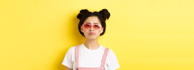 Sommer- und modekonzept gelangweiltes asiatisches teenager-mädchen mit sonnenbrille, das launisch auf gelbem rücken steht