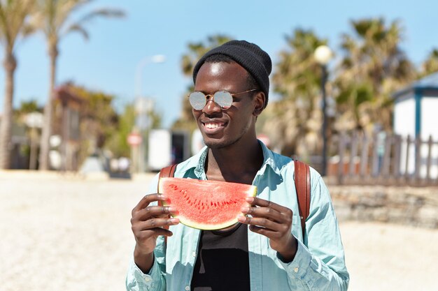 Sommer, Urlaub, Ferien und Lifestyle. Sorgloser glücklicher junger dunkelhäutiger männlicher Reisender, der kleines Picknick mit Freunden am Meer hat und saftige köstliche Wassermelone isst
