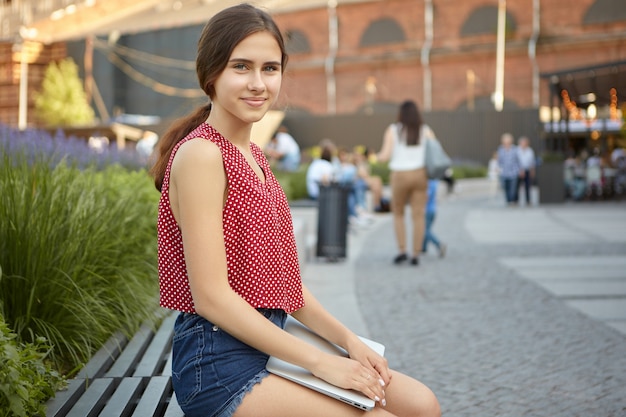 Sommeransicht im Freien der schönen positiven jungen Frau im gepunkteten roten Oberteil und in den Jeansshorts unter Verwendung des tragbaren elektronischen Geräts im Park, schauend und lächelnd. Moderne Technologie und Kommunikation