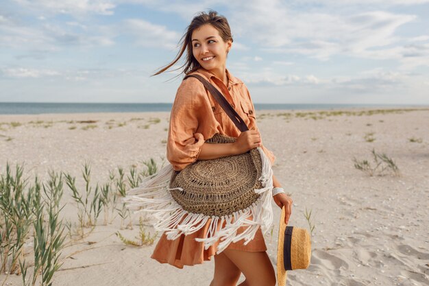 Sommerbild der schönen brünetten Frau im trendigen Leinenkleid, das herumspringt und herumalbert und Strohsack hält. Hübsches schlankes Mädchen, das Wochenenden nahe Ozean genießt.