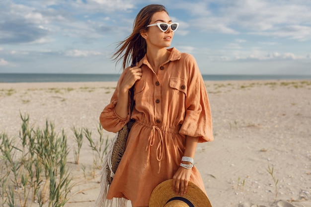 Sommerbild der schönen brünetten Frau im trendigen Leinenkleid, Strohsack haltend. Hübsches schlankes Mädchen, das Wochenenden nahe Ozean genießt.