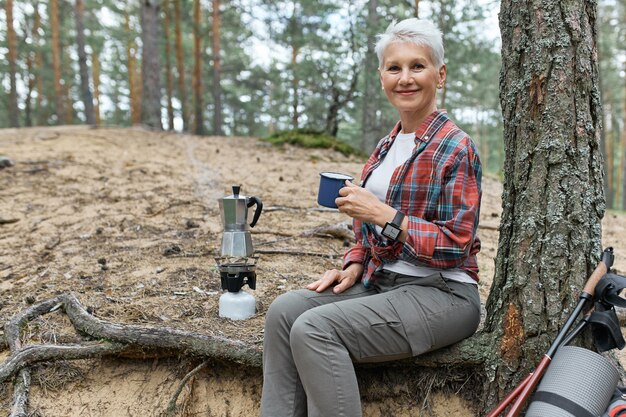 Sommerbild im Freien der fröhlichen Frau mittleren Alters in der Aktivkleidung, die unter Baum mit Campingausrüstung und Kessel auf Gasbrenner entspannt, Becher hält, frischen Tee genießt, Ruhe beim Wandern allein hat