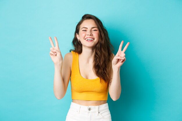 Sommerferien und Emotionen Konzept. Glückliches schönes Mädchen, das Friedens-V-Zeichen zeigt und Spaß hat, in die Kamera lacht und lächelt und vor blauem Hintergrund steht.