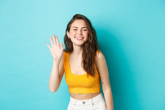 Sommerferien und Emotionen Konzept. Schöne freundliche Frau mit perfektem Körper, winkender Hand und sagt Hallo, grüßt dich und steht auf blauem Hintergrund