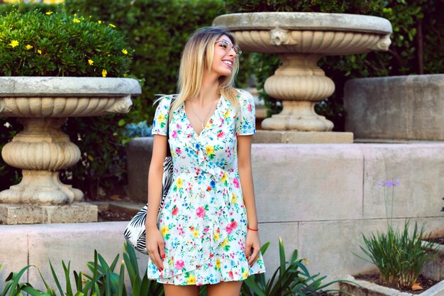 Sommerlebensstilporträt der glücklichen prächtigen blonden stilvollen Frau, die leichtes elegantes trendiges Blumentee-Kleid trägt und im alten europäischen Park, Reisestimmung aufwirft.