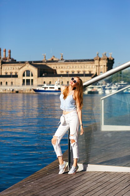 Sommerlebensstilporträt der hübschen blonden Frau reisen allein in Barcelona, schöne Architektur und Meerblick, trendiger Straßenstilblick, Urlaub, Freude, Reisender, Erntedach und Denim.