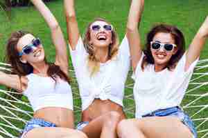 Kostenloses Foto sommerlebensstilporträt von baumfrauen, die verrückt werden, schreien, lachen, spaß zusammen haben, auf hängematte springen. trägt weiße tops und sonnenbrillen, bereit für party, freude, spaß.
