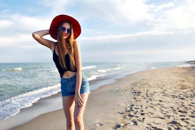 Sommermodeporträt der jungen Frau, die allein in der Nähe des Ozeans geht, Urlaub am Strand, allein reisen, Vintage Hut Sonnenbrille und Jeansshorts tragen, schlanker Körper, Sonnenaufgang, gesunder Lebensstil.