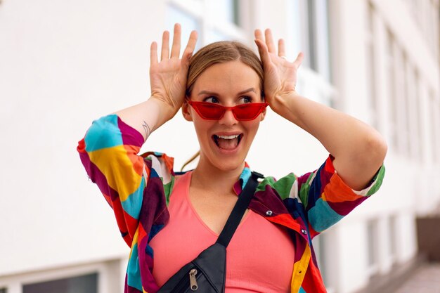 Sommernahes sonniges Porträt der hübschen blonden Frau, die helle Freizeitkleidung und Sonnenbrille trägt und lustige Gesichter macht.