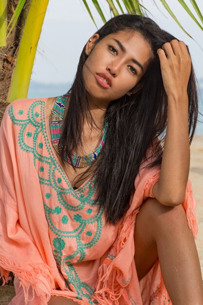 Sommerporträt der lächelnden hübschen asiatischen Frau in der rosa stilvollen Strandkleidung, die auf Sand nahe Palme, blauer Ozean sitzt. Schmuck, Armband und Halskette.