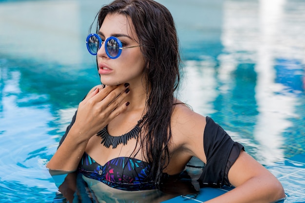 Sommerporträt des schönen sexy Modells in der schwarzen Badebekleidung und in der stilvollen Halskette, die im Pool aufwirft. Urlaub und tropische Stimmung.