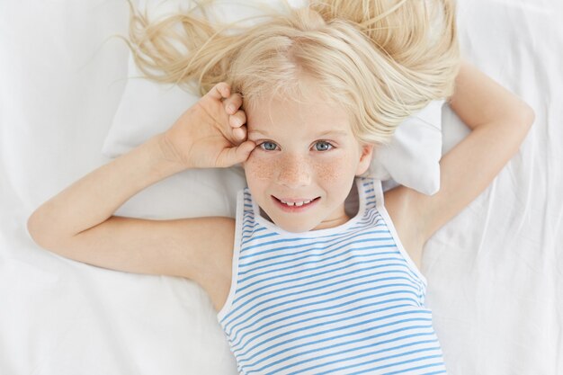 Sommersprossiges blauäugiges Mädchen mit blonden Haaren, gestreiftem T-Shirt, mit entzückendem Ausdruck schauend, während auf weißen Bettwäsche liegend. Hübsches kleines weibliches Kind, das guten Morgen im Bett genießt.