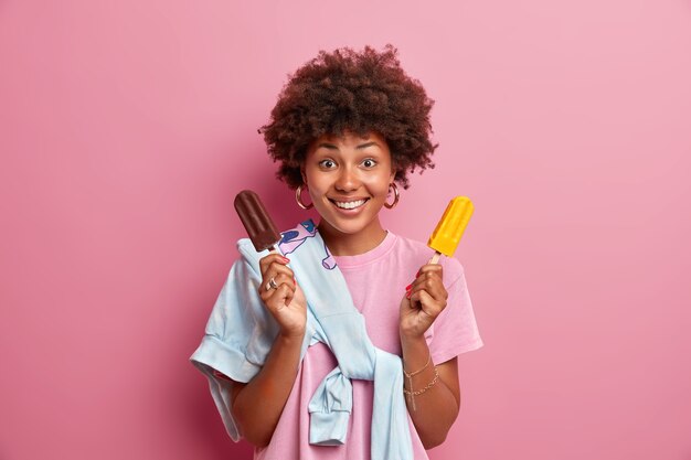 Sommerzeitkonzept. Frohe positive Frau mit Afro-Frisur hält leckeres gefrorenes Eis, genießt es, leckeres kaltes Dessert zu essen, lässig gekleidet, posiert