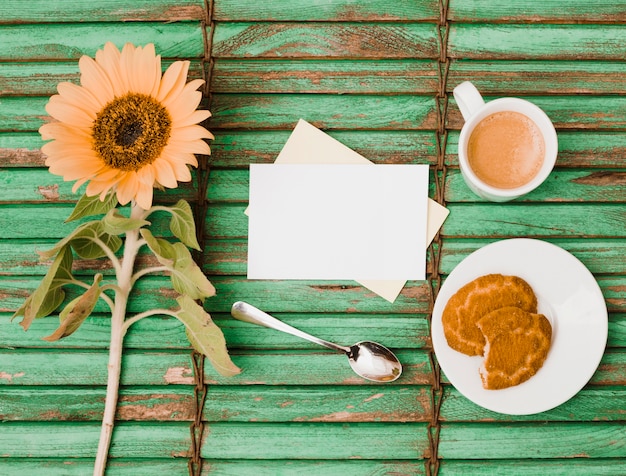 Kostenloses Foto sonnenblume; löffel; leere karten; gegessene plätzchen und kaffeetasse auf grünem hölzernem hintergrund